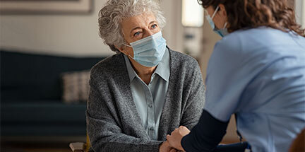 An elderly woman wearing a face mask holds hands with a care worker also wearing a face mask