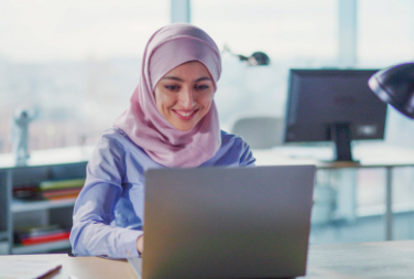 A woman wearing a hijab works on a laptop with another desk and computer in the background