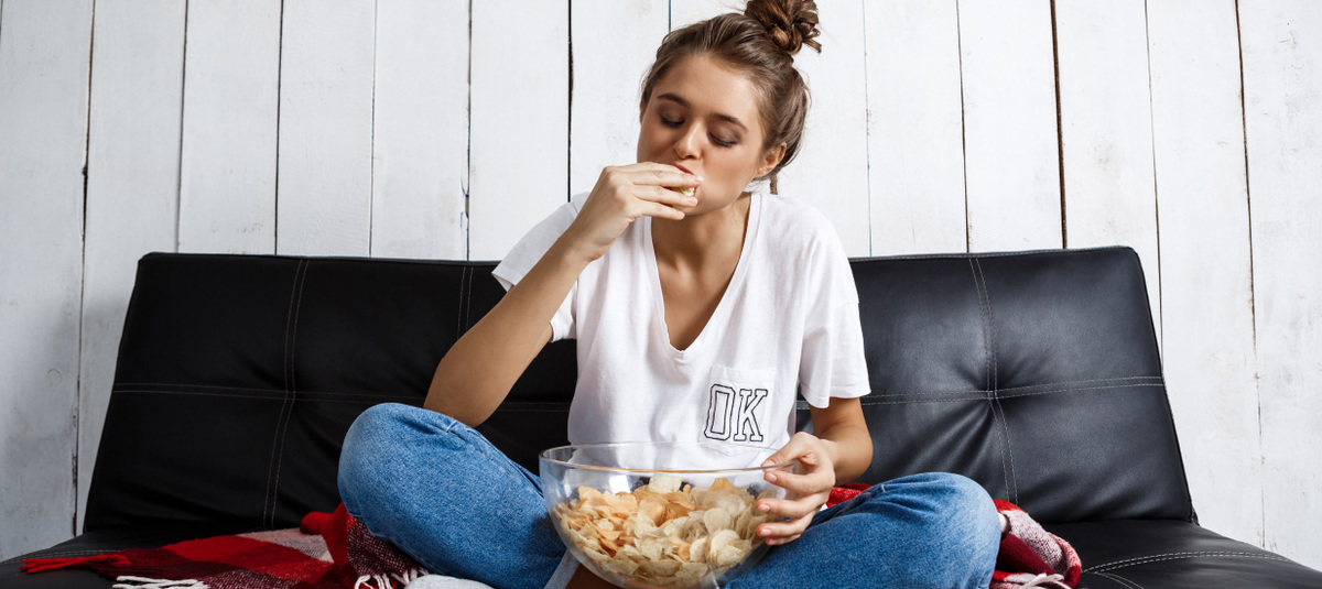 A woman sits on a couch and emotionaly eats by stuffing her mouth full of potato chips.