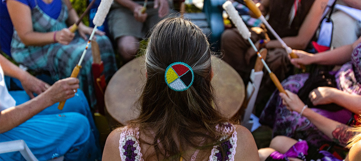 The back of a person with long hair, pulled back with a red, yellow, black and white hair clip faces an audience or crowd of people beating drums